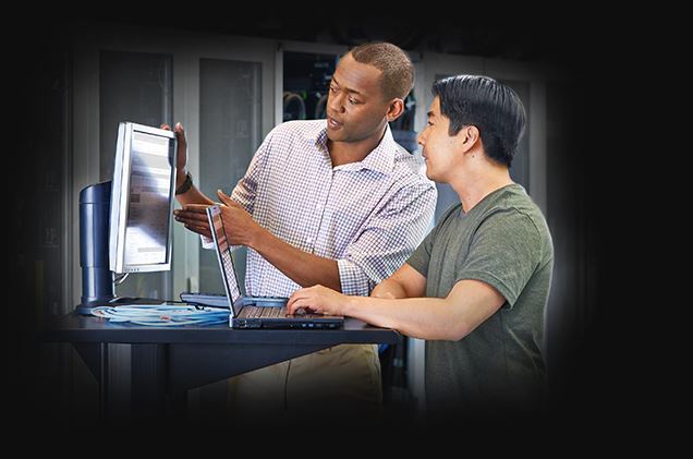 two men talking while using a laptop and looking at a computer screen