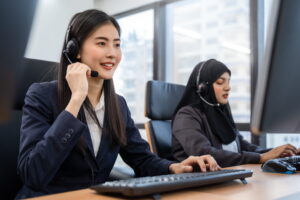 Two customer service agents wearing headsets handle calls at a shared workstation in a modern, well-lit office