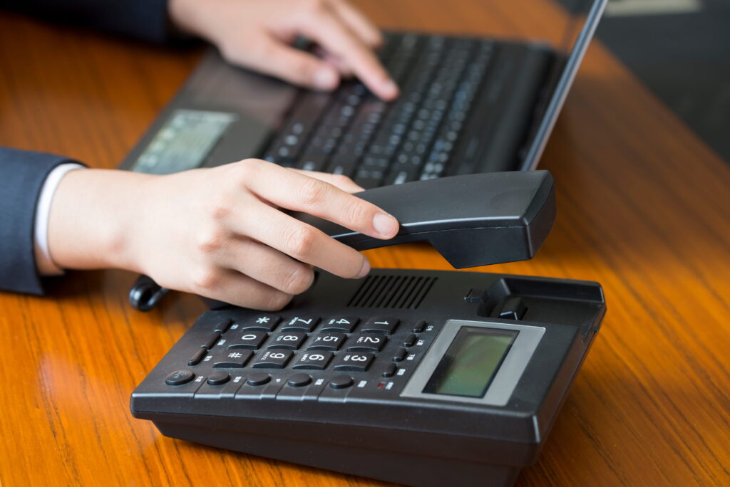A business professional in a dark suit lifts the handset of a black office desk phone while working on a laptop at a wooden desk