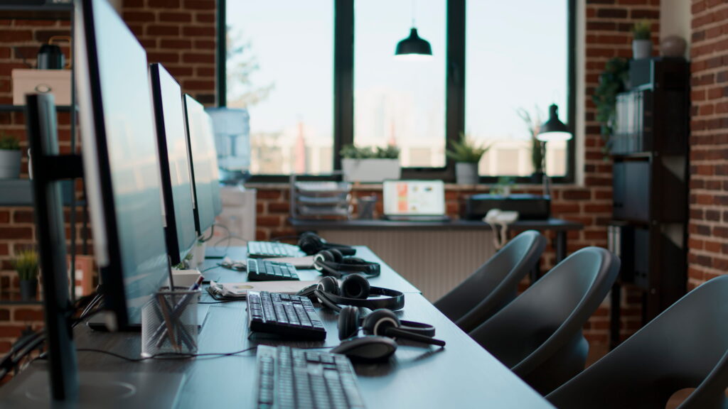 A row of computer monitors, keyboards, and communication headsets sits empty along a shared desk in a modern open-plan office with exposed brick walls and large windows
