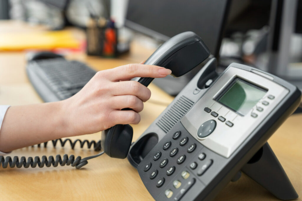 A person's hand picking up the receiver of a black VoIP phone in an office.