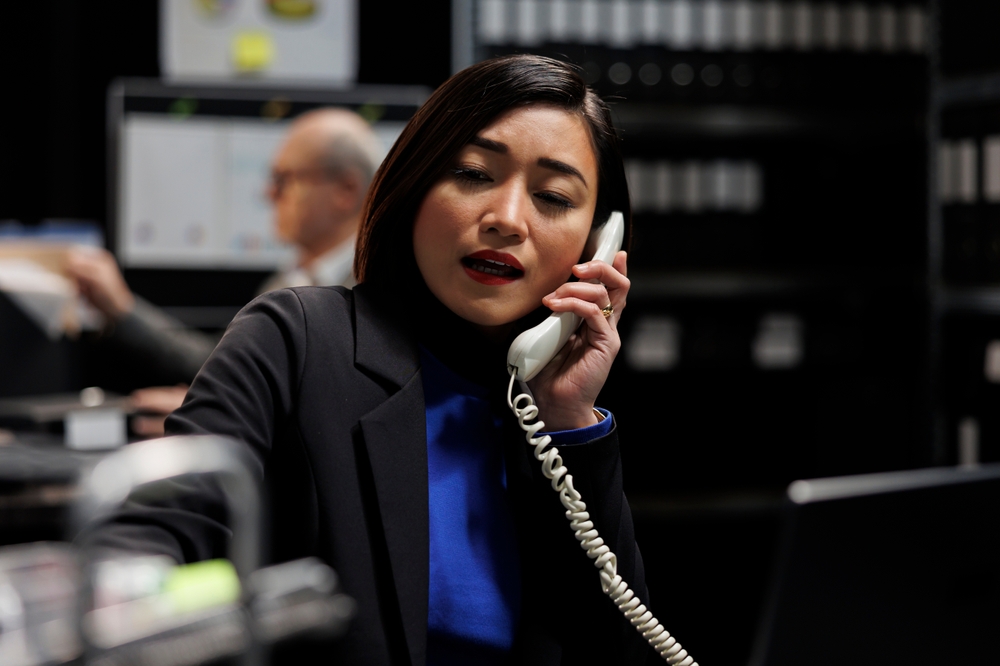 A female professional in a black blazer speaking on a VoIP at her office desk