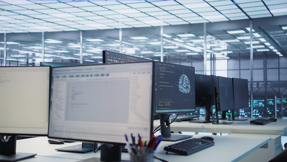 Multiple computer monitors displaying code and data graphics on desks inside a modern server room or data center.